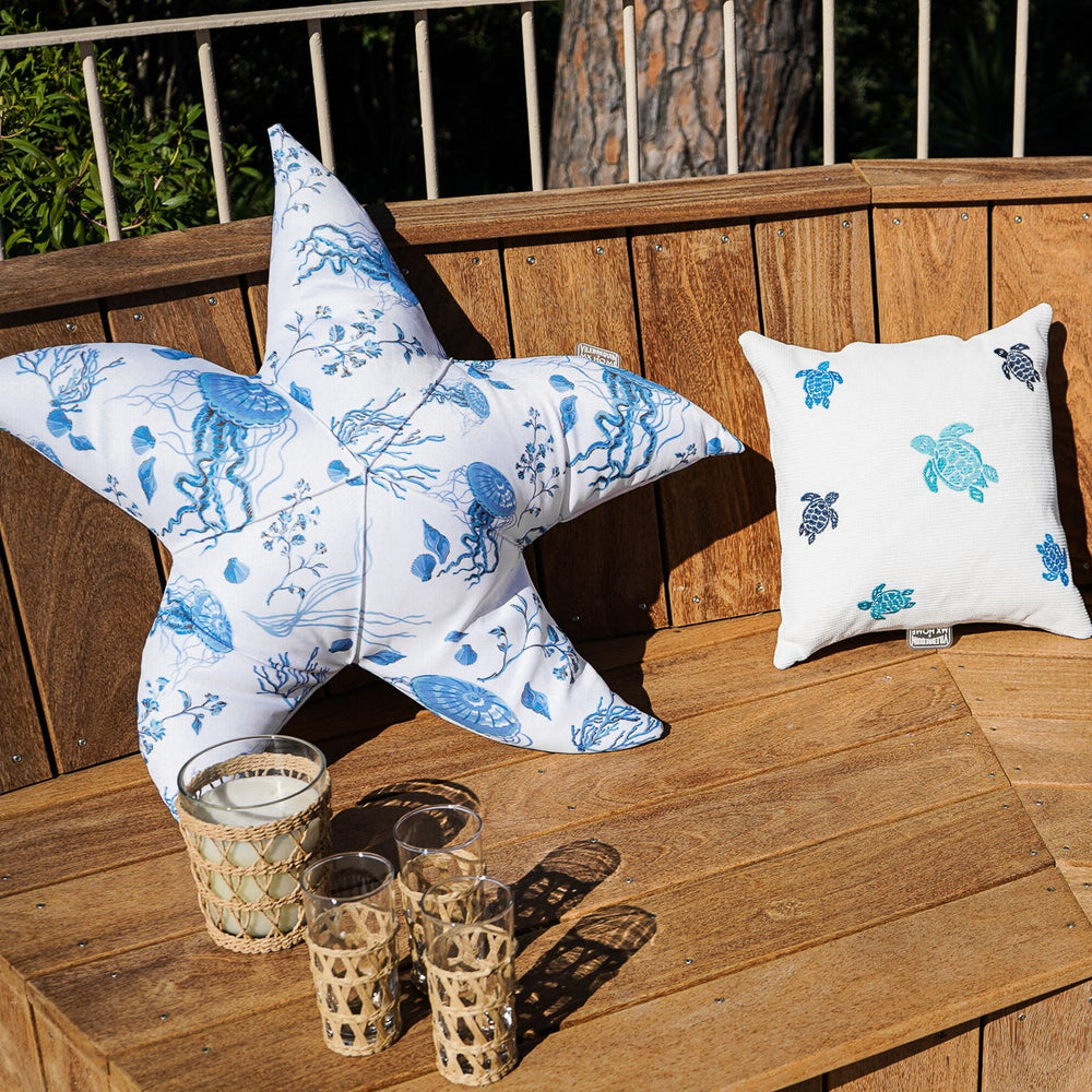 Scene with two white Vilebrequin cushions, one embroidered with multicolored turtles and the other with an integrated blue jellyfish motif, in an elegant outdoor setting.