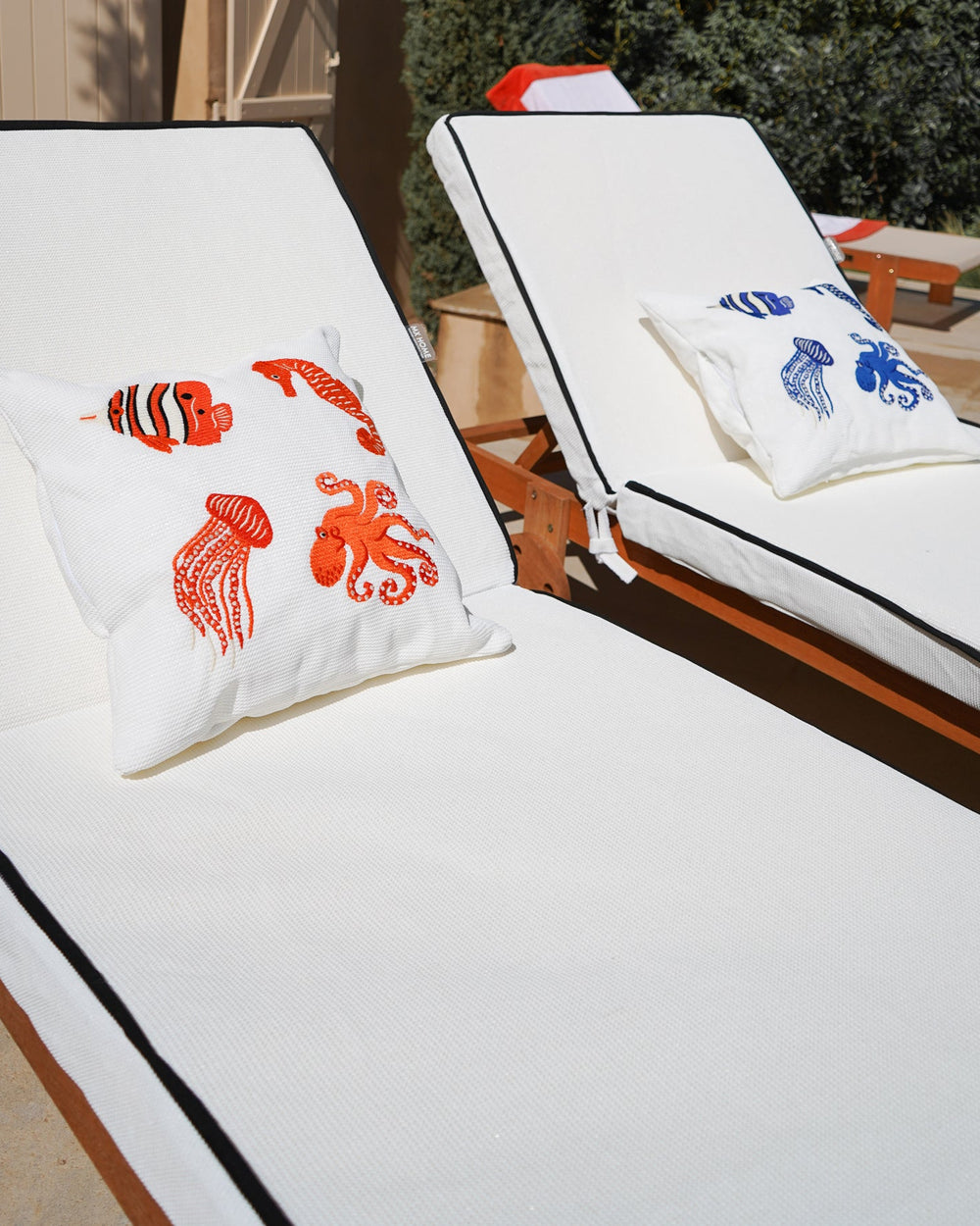 Série de coussins brodés à motifs marins posés côte à côte sur nos matelas blancs à passepoils noirs sur une terrasse.